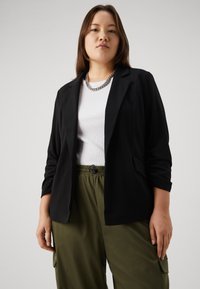 Černý blazer s hladkou texturou, zapínání na jeden knoflík a dvěma předními kapsami, v kombinaci s bílým žebrovaným tričkem a olivově zelenými kalhotami.