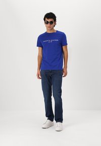 Blaues Baumwoll-T-Shirt mit weißem Logo-Text, kombiniert mit dunklen Jeans und weißen Turnschuhen. Sonnenbrillen runden das Outfit ab.