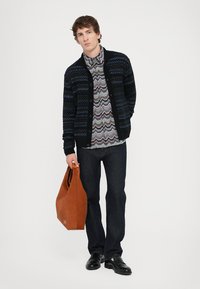 Adīts melns kardigans ar ziliem rakstiem, daudzkrāsains zigzaga krekls, tumšas džinsi un brūna ādas somiņa, apgādāts ar melnām kurpēm.