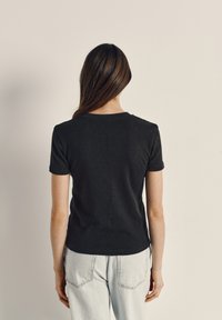 Moteris ilgais rudais plaukais stovi veidu į paprastą kreminės spalvos sieną, dėvi juodą trumpomis rankovėmis marškinėlį ir šviesiai mėlynas džinsines kelnes.