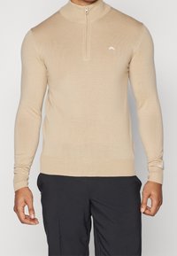 Beige zip-up tröja i mjukt stickat material, med ribbad krage, manschetter och fåll, samt en liten vit logotyp på bröstet.