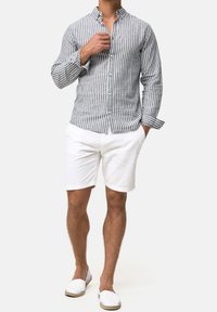 Man die een grijs-wit gestreept overhemd met lange mouwen draagt, witte shorts en witte instapschoenen, staand met één hand in zijn zak.