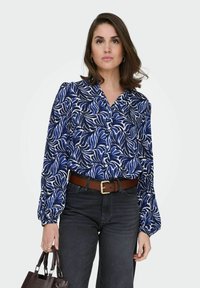 Femme portant une blouse à motifs de feuilles bleu et blanc avec des manches bouffantes, un jean noir, une ceinture marron, et tenant un sac à main marron foncé.