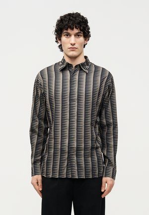 Ung mann med krøllete mørkt hår iført en langermet skjorte med vertikale svarte og beige sikksakstriper, stående foran en ensfarget bakgrunn.