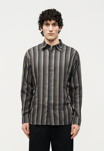 Joven con cabello rizado y oscuro que lleva una camisa de manga larga con rayas en zigzag verticales negras y beige, de pie contra un fondo liso.