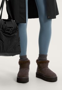 Barna velúr bokacsizmák műszőrme díszítéssel, lapos gumitalppal, szürke bordázott zoknikkal és kék leggings-szel párosítva. Fekete táska a közelben.