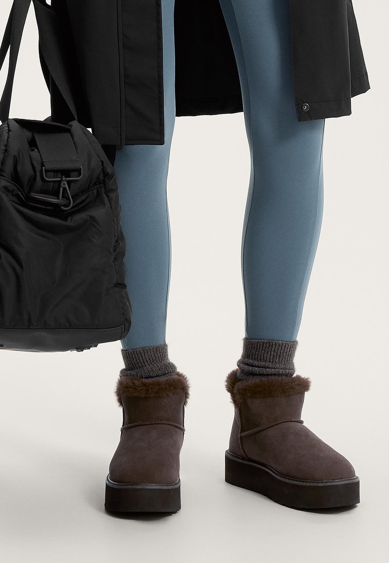 Barna velúr bokacsizmák műszőrme díszítéssel, lapos gumitalppal, szürke bordázott zoknikkal és kék leggings-szel párosítva. Fekete táska a közelben.