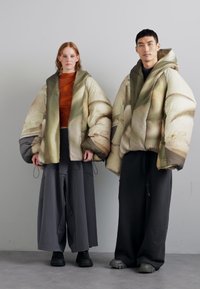 Padded jackets with a textured, abstract pattern in muted earth tones, worn over loose, dark trousers; layered, oversized design with hoods.