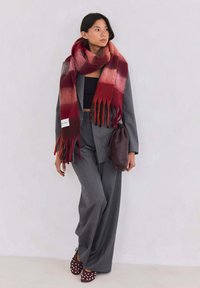 Écharpe dégradée rouge et rose avec des franges, portée sur un costume noir. Sac marron et chaussures bordeaux avec des accents blancs complètent la tenue.