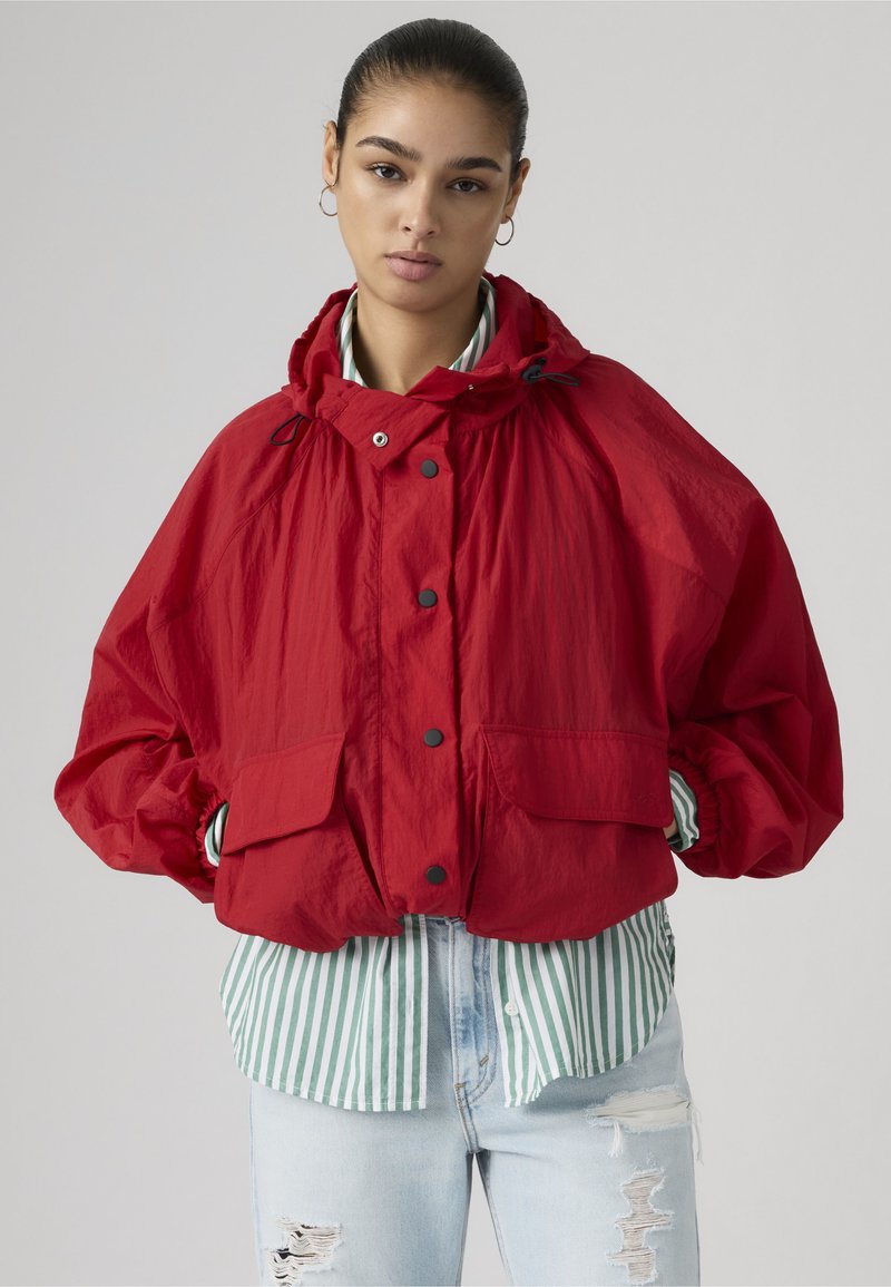 Junge Frau mit dunklen Haaren, die eine lockere rote Jacke über einem grün-gestreiften Hemd und zerrissene hellblaue Jeans trägt, steht vor einem einfarbigen Hintergrund.