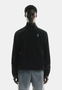 Black windbreaker with a zipper collar, featuring a logo on the left chest. Lightweight material and elastic cuffs. Worn over light gray shorts.