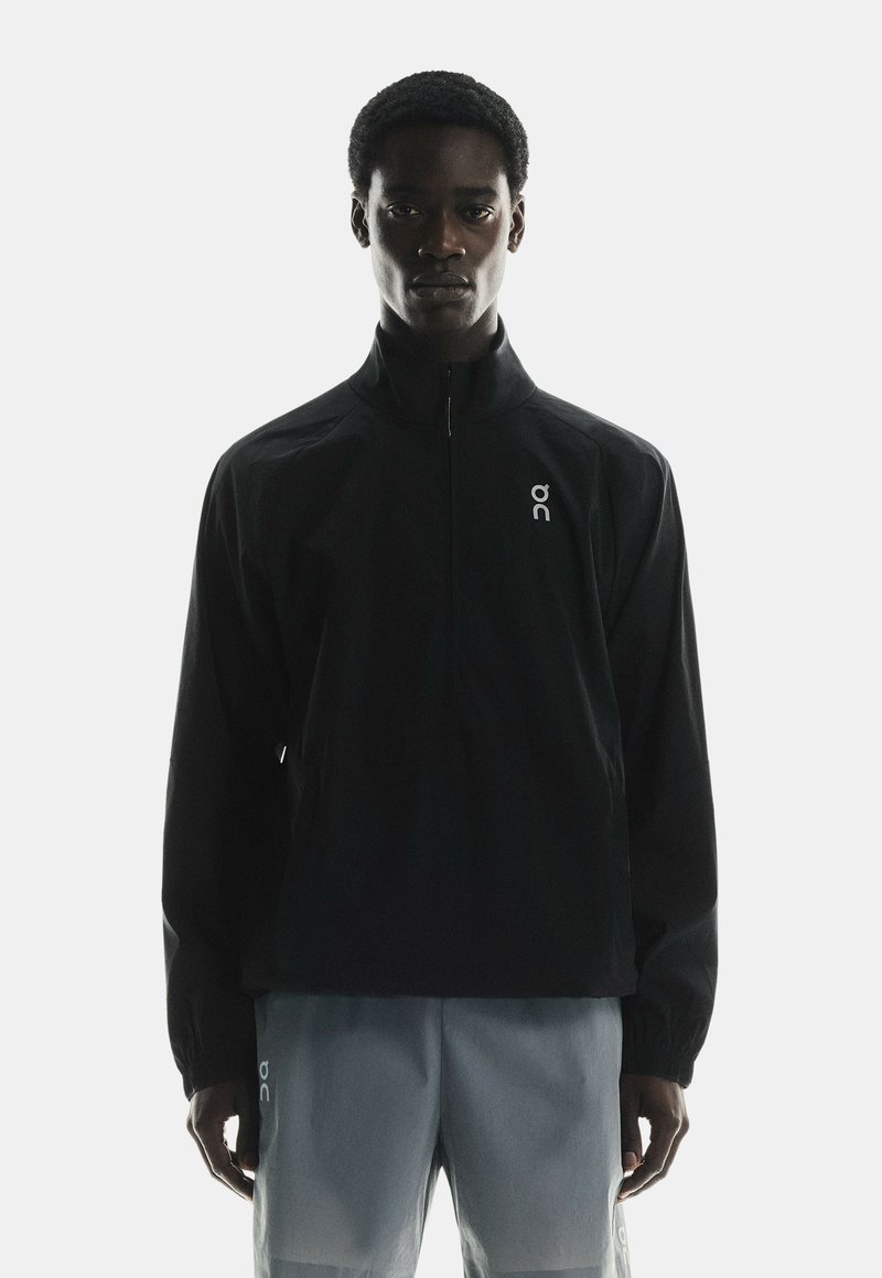 Black windbreaker with a zipper collar, featuring a logo on the left chest. Lightweight material and elastic cuffs. Worn over light gray shorts.