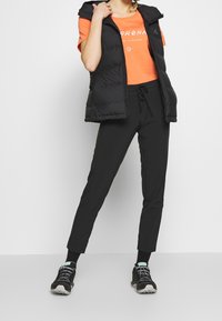 Svart puffväst med huva, orange grafisk t-shirt, svarta dragsnörningsbyxor och svarta atletiska skor med ljusblå accenter.