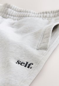 Heledad hallid dressipüksid elastse vöökohaga ja musta tikandiga tekstiga "self." ees tasku lähedal.