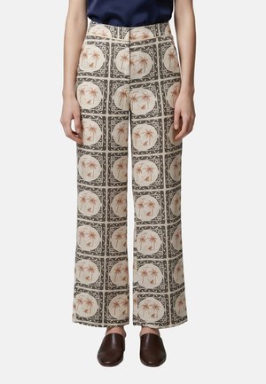 Pantalon taille haute à jambes larges avec motif carré répétitif présentant des palmiers et des huttes dans des tons terre, porté avec des chaussures foncées et un haut bleu marine.