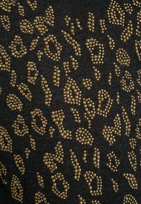 Černá tkanina s texturovaným povrchem, na které jsou uspořádány zlaté kovové nýty ve vzoru leopardího skvrnitého. 