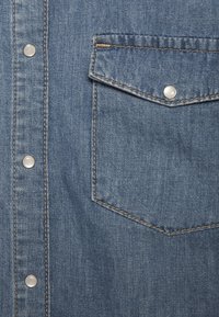 Chemise en denim avec poche avant à rabat sécurisée par un bouton-pression métallique et rangée verticale de boutons-pression métalliques le long de la patte de boutonnage.