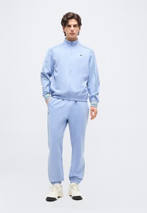 Joven de pie con las manos en los bolsillos, vistiendo un chándal azul claro y zapatillas blancas frente a un fondo blanco liso.