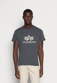 Graues Baumwoll-T-Shirt mit Rundhalsausschnitt, das ein großes wei�ßes Logo und den Text "ALPHA INDUSTRIES" auf der Vorderseite trägt. Mit kurzen Ärmeln.