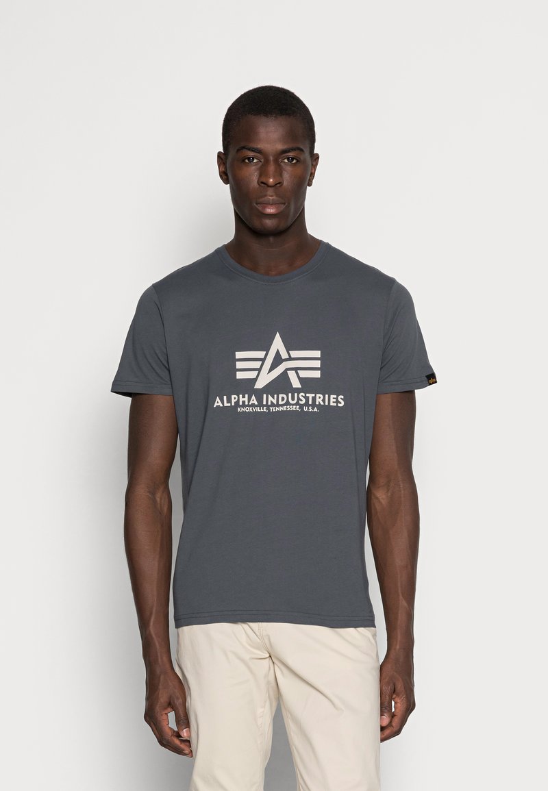 Graues Baumwoll-T-Shirt mit Rundhalsausschnitt, das ein großes weißes Logo und den Text "ALPHA INDUSTRIES" auf der Vorderseite trägt. Mit kurzen Ärmeln.