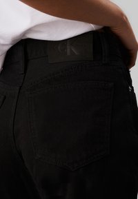Jeans de mezclilla negra con un bolsillo rectangular y una etiqueta de cuero negro con la inscripción "Calvin Klein Jeans" en la cintura, con detalles de costura sutiles.