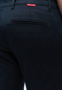Nærbillede af lommen bagpå sorte bukser med en rød "JOOP! JEANS" label over den vandrette slidslomme.