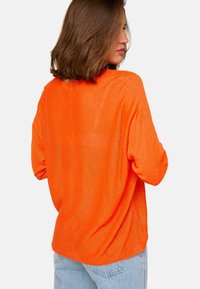 Femme portant un pull orange vif, ample et à manches longues, associé à un jean en denim bleu clair, de profil avec le dos visible.