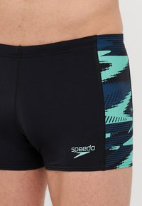 Černé plavky s bočním panelem, který má modro-aqua grafický vzor. Vyrobeny z hladkého materiálu s přiléhavým střihem.