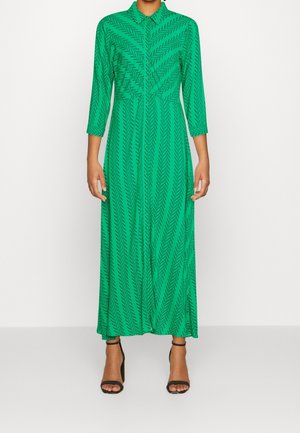 Femme portant une robe longue verte à manches longues avec un motif chevron et des sandales noires à talons hauts, debout devant un fond uni.