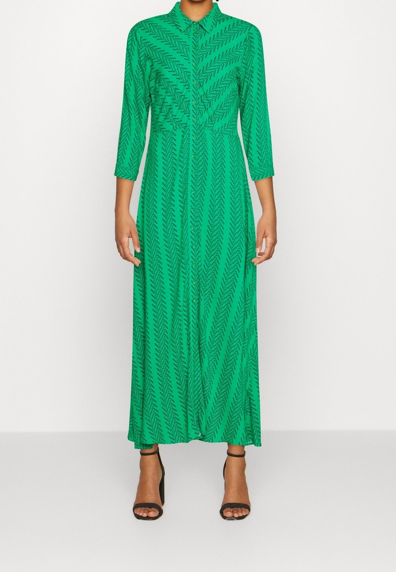 Femme portant une robe longue verte à manches longues avec un motif chevron et des sandales noires à talons hauts, debout devant un fond uni.