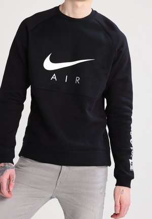 Osoba ubrana w czarną bluzę Nike Air z białym logo i jasnoszare spodnie, stojąca na jednolitym jasnym tle.