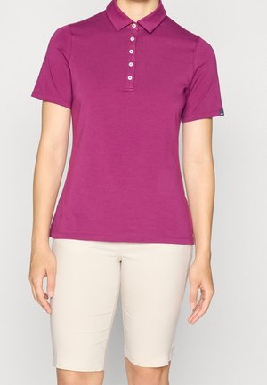 Kvinde iført en kortærmet magenta polo med hvide knapper og beige knælange shorts, stående mod en ensfarvet baggrund.
