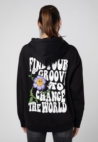 Zwarte hoodie met een grote witte grafische tekst die zegt "VIND JOUW GROOVE OM DE WERELD TE VERANDEREN" met een lachende paarse bloem en groene bladeren.