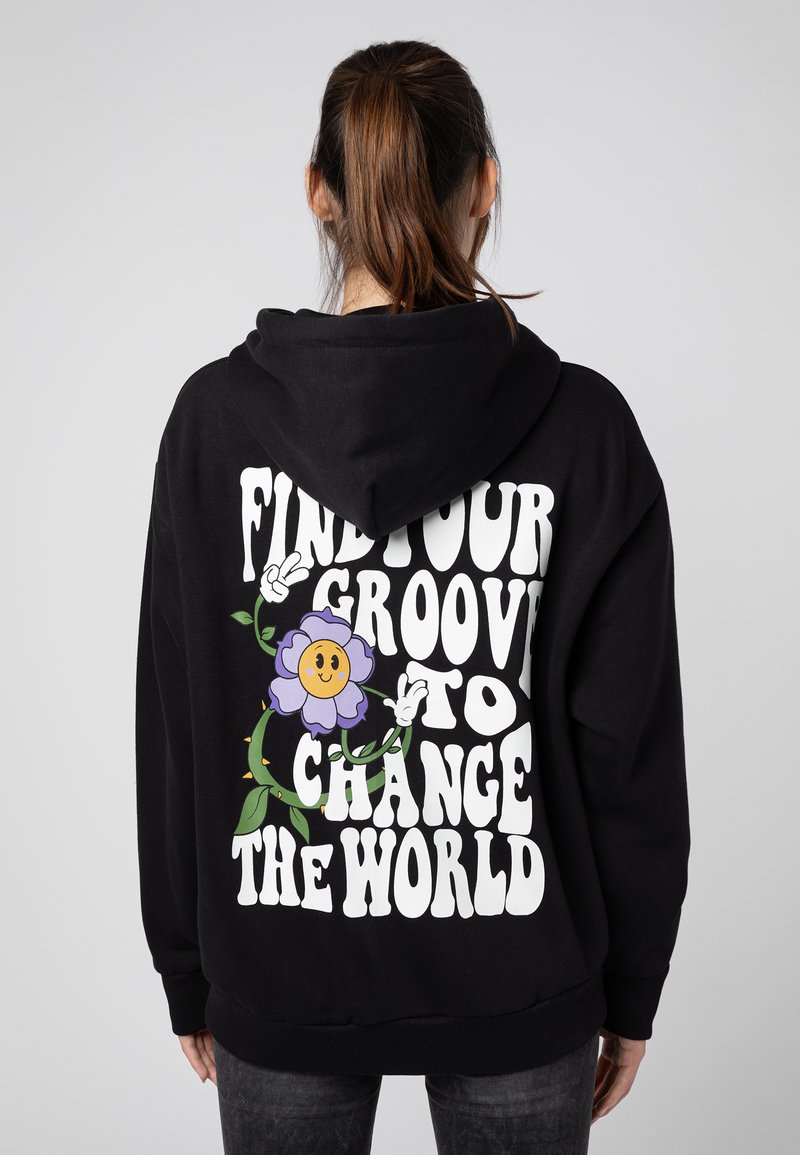 Zwarte hoodie met een grote witte grafische tekst die zegt "VIND JOUW GROOVE OM DE WERELD TE VERANDEREN" met een lachende paarse bloem en groene bladeren.