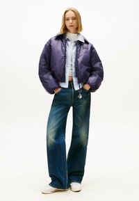 Giacca a vento viola dal taglio corto e colletto ampio, abbinata a jeans in denim blu a gamba larga. Gli accessori includono un charm sui jeans.