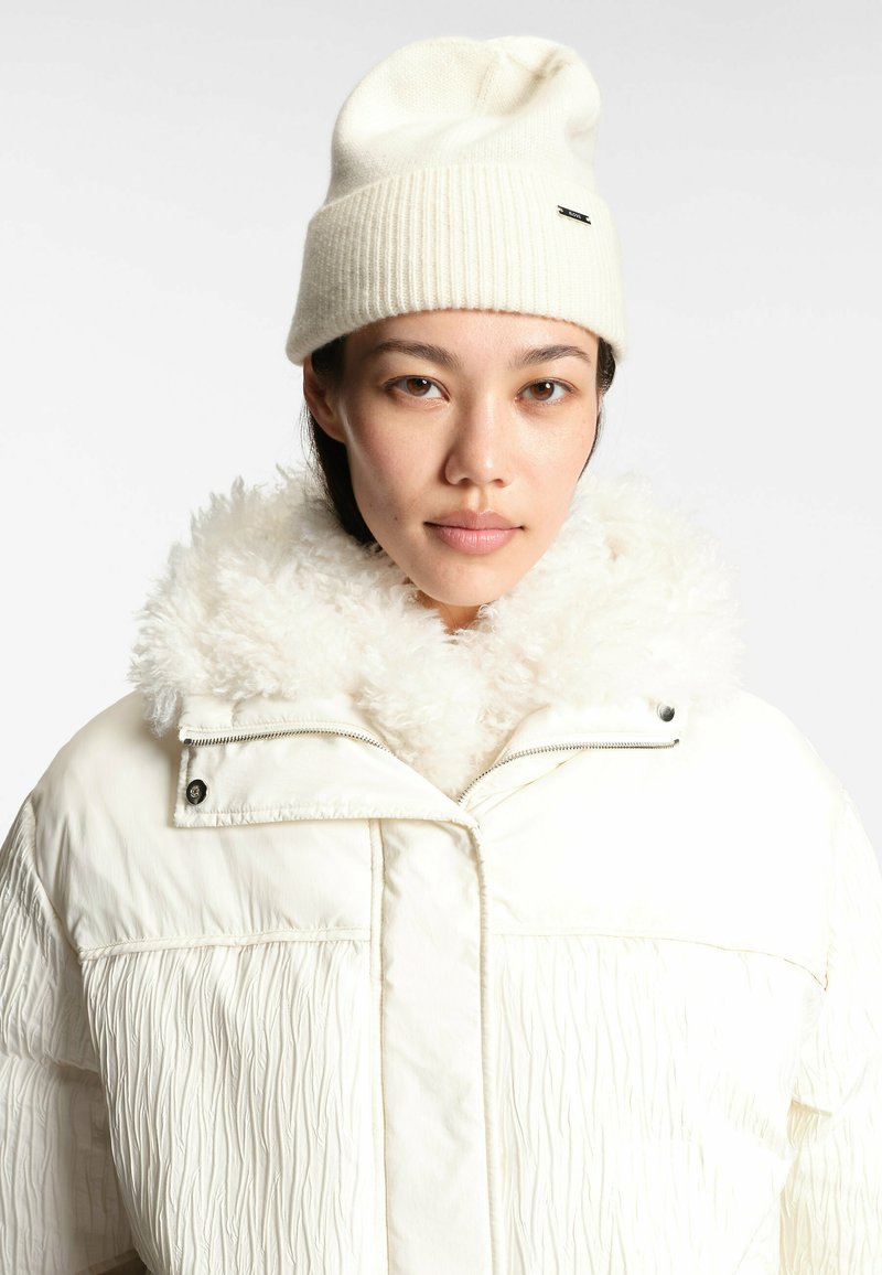 Vit pufferjacka med texturerad tyg och krage av fuskpäls, kombinerad med en krämfärgad stickad mössa. Dragkedjedetalj synlig på jackan.