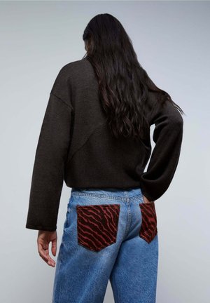 Suéter oversized marrón oscuro combinado con jeans de mezclilla azul claro que cuentan con bolsillos de rayas de tigre rojas. Textura suave y corte relajado.