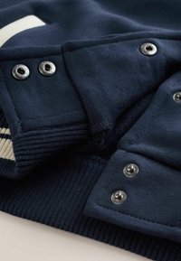 Mørkeblå sweatshirt med ribstrikkede manchetter, udstyret med sølvfarvede trykknapper og en glat tekstur. Delvist synlig cremefarvet stribedetalje.