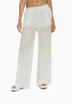 Kobieta ubrana w luźne, przezroczyste białe spodnie o szerokich nogawkach oraz białe platformy z odkrytymi palcami, stojąca na tle białego tła.