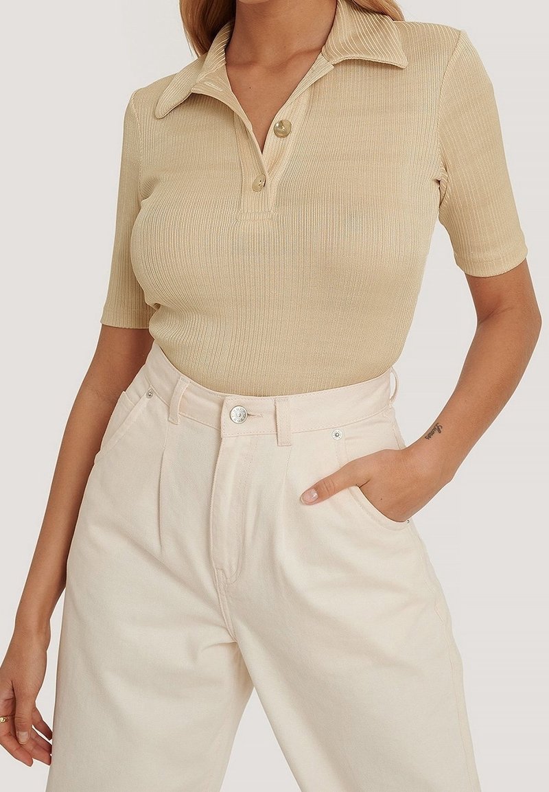 Femme portant un polo beige à manches courtes côtelé, rentré dans un pantalon large crème taille haute, une main dans la poche.
