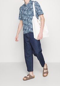 Camicia a maniche corte, blu con motivo tropicale abbinata a jeans larghi blu scuro e sandali marroni a scivolo. Tiene una borsa tote bianca.