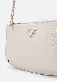 Borsa beige con finitura testurizzata, caratterizzata da una piastra con logo dorato a forma triangolare e una breve tracolla dorata a catena. Dettaglio con chiusura a zip visibile.