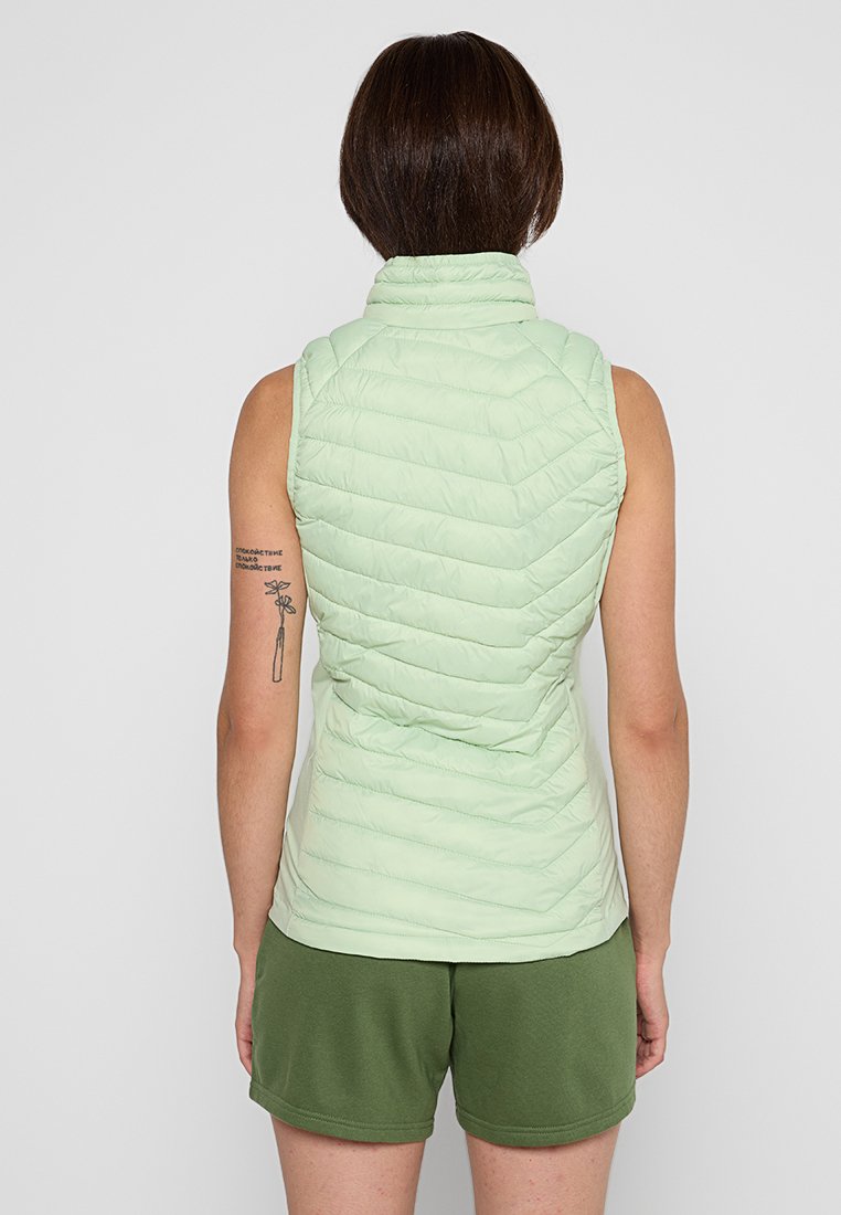 Femme aux cheveux bruns courts portant un gilet sans manches matelassé vert clair et un short vert, se tenant de dos face à l'appareil photo.