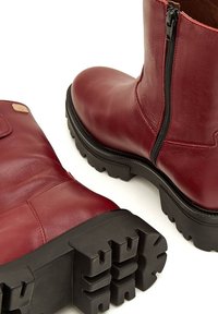 Botas de tobillo de cuero burdeos con cremallera lateral, suela de goma negra texturizada y puntera reforzada. Presenta un pequeño logo en relieve en el costado.