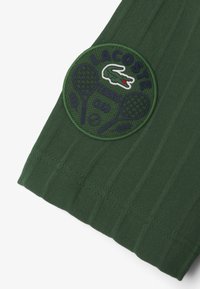 Grön texturerad tyg med en rund patch med "LACOSTE TENNIS CLUB" och en krokodil-logotyp, dekorerad med tennisracketer och blad.