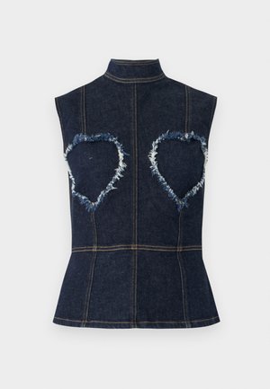 Nő sötét farmer ujjatlan felsőben, amelyen szív alakú, foszlott foltok vannak, hosszú bézs szoknyában, színes nyomtatott grafikákkal, és fekete magas sarkú cipőben.