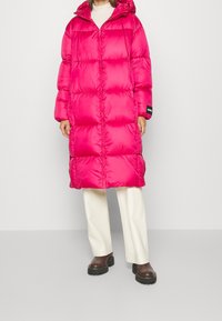 Abrigo acolchado rosa con capucha, presenta un diseño acolchonado y cremallera frontal. Se lleva con pantalones crema y botas negras gruesas.