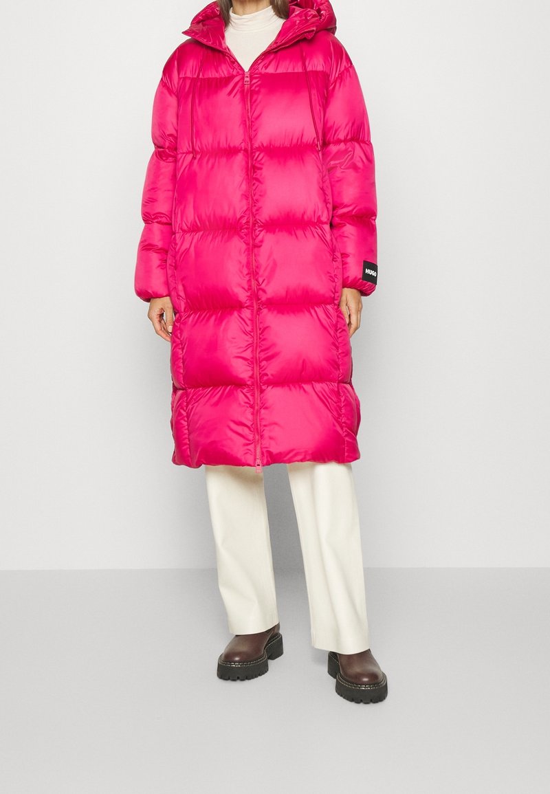 Abrigo acolchado rosa con capucha, presenta un diseño acolchonado y cremallera frontal. Se lleva con pantalones crema y botas negras gruesas.