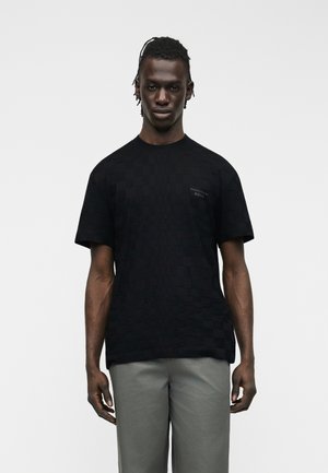 Άνδρας που στέκεται με το πρόσωπο μπροστά, φοράει μαύρο καρό κοντομάνικο πουκάμισο και ανοιχτό γκρι παντελόνι μπροστά σε μονόχρωμο φόντο.