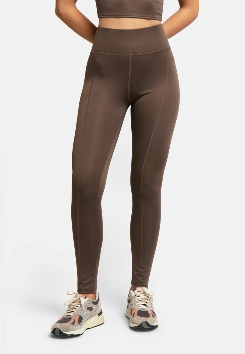 Leggings marron taille haute portés avec des chaussures de course multicolores beige, pêche et noir sur une personne debout les mains dans le dos.
